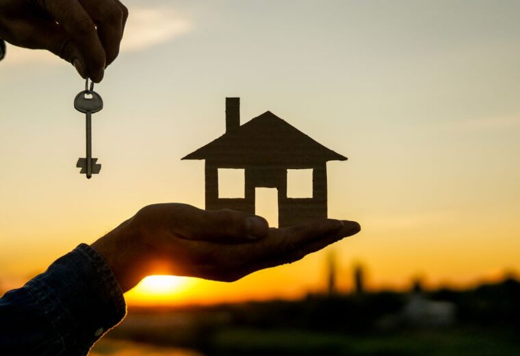 A hand holding a small wooden house with a key in the other hand, in front of a sunset - Express Capital Financing