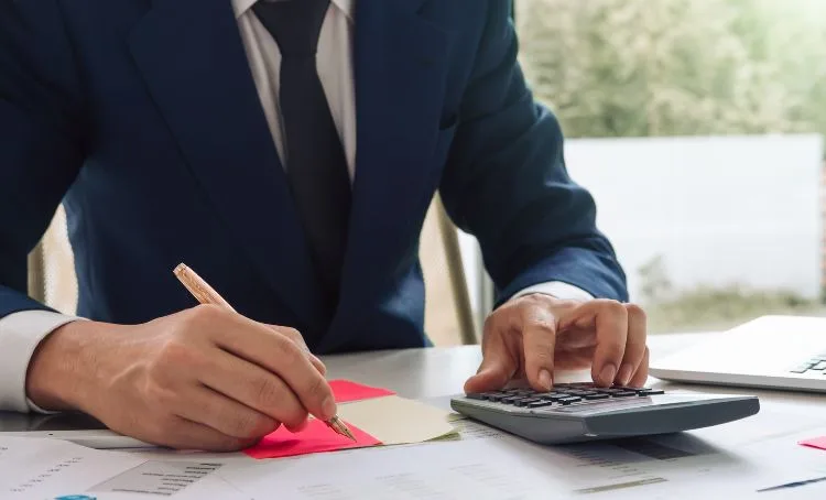 A man in a suit sitting at a desk writing with one hand while sung a calculator with the other - Express Capital Financing